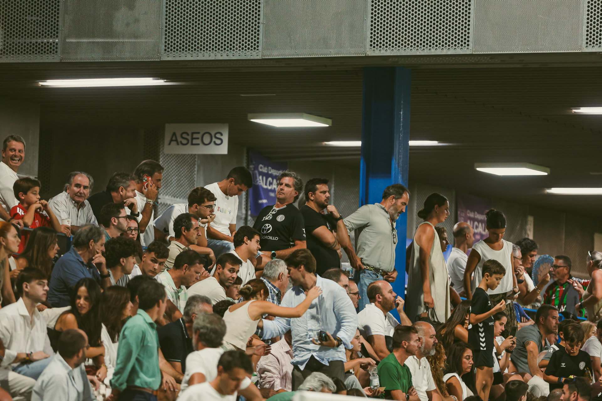 La tribuna llena de noche siguiendo el partido